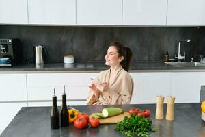 ritratto di donna nel accappatoio seduta nel cucina con smartphone, cucinando cena, Guardando ricetta su sociale media, video lezione Come per preparare pasto foto