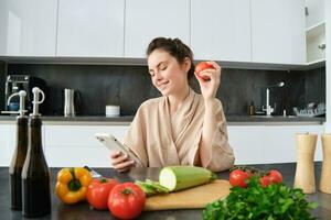 ritratto di giovane donna ordinazione drogheria su smartphone app, Tenere pomodoro, seduta vicino chopping tavola con verdure. ragazza guardare per ricetta in linea, utilizzando mobile Telefono foto