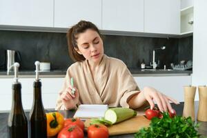 ritratto di donna cucinando nel il cucina, seduta nel davanti di la verdura, pomodori zucchine e prezzemolo, fabbricazione elenco di drogheria, scrittura giù ricetta, indossare accappatoio foto