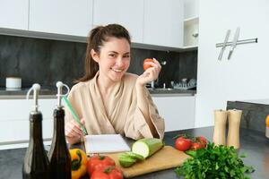 ritratto di bellissimo sorridente donna, scrittura sua salutare menù, mangiare pomodoro mentre cucinando, fabbricazione drogheria elenco, seduta nel il cucina foto