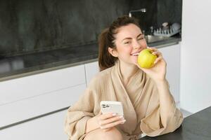 vicino su ritratto di contento giovane donna nel accappatoio, seduta nel il cucina e utilizzando mobile Telefono, Tenere un mela, ordine frutta e verdure in linea, utilizzando smartphone App per drogheria consegna foto