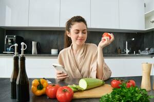 giovane donna ordini drogheria su mobile app. ragazza nel accappatoio si siede nel il cucina con la verdura, guardare per ricetta per cucinare cena, utilizzando smartphone applicazione foto