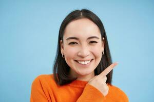 vicino su ritratto di sorridente asiatico donna, puntamento dito Giusto, mostrando annuncio, copia spazio striscione, in piedi al di sopra di blu sfondo foto