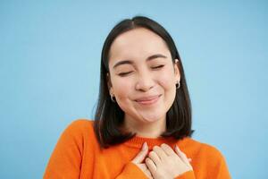 vicino su di bello asiatico donna, detiene mani su cuore, sognare ad occhi aperti, pensiero di qualcosa felice, in piedi al di sopra di blu sfondo foto