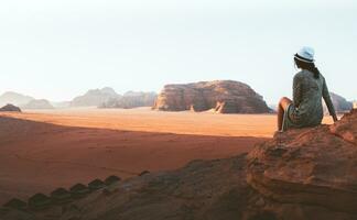 donna turista nel vestito sedersi su scogliera a punto di vista su tramonto nel wadi Rum deserto - valle wadi saabit. Giordania Esplorare concetto foto