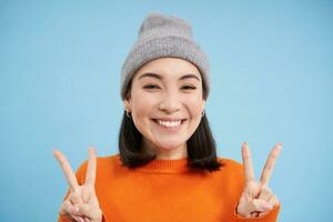 vicino su ritratto di positivo e carino asiatico ragazza nel berretto, sorrisi e Spettacoli pace, kawaii cartello blu sfondo foto