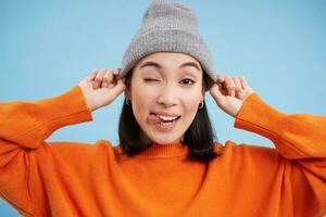 vicino su ritratto di asiatico femmina modello mette su cappello e strizza l'occhio a voi, guardare felice, andando fuori, in piedi al di sopra di blu sfondo foto