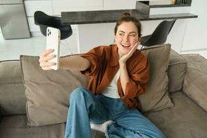 foto di sorridente giovane donna seduta su divano, in posa su divano nel vivente camera, assunzione selfie per sociale media