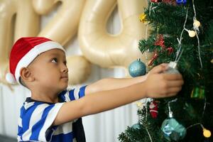 allegro Natale 2024 concetto asiatico ragazzo decora Natale albero con colorato Natale palle. foto