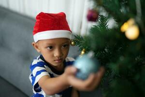 allegro Natale 2024 concetto asiatico ragazzo decora Natale albero con colorato Natale palle. foto