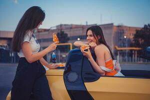 giovane ragazze nel casuale attrezzatura siamo sorridente, mangiare Pizza, in posa nel giallo auto cabrio con francese patatine fritte e bibita nel bicchiere bottiglia su suo tronco. veloce cibo foto