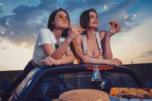 Due donne mangiare Pizza, sorridente, in posa nel giallo auto cabrio con francese patatine fritte, cappello e bibita nel bicchiere bottiglia su tronco. veloce cibo. vicino su, finto su foto
