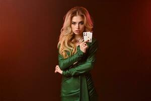 bionda ragazza nel verde elegante vestito e gioielleria. mani piegato, mostrando Due assi, in posa contro Marrone studio sfondo. poker, casinò. avvicinamento foto