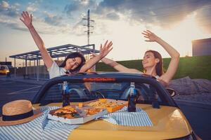 giovane ragazze sorridente, sollevato su mani, in posa nel giallo auto con francese patatine fritte, Pizza e bibita nel bicchiere bottiglie su suo tronco. veloce cibo. copia spazio foto