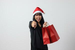 ritratto di allegro asiatico donna con rosso Santa cappello sensazione contento mentre puntamento per il lato. eccitato ragazza Tenere shopping Borsa. nuovo anno e Natale concetto. isolato Immagine su bianca sfondo foto