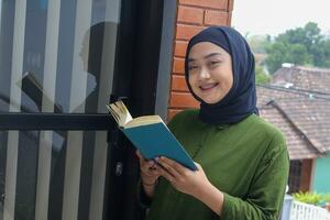 ritratto di attraente asiatico hijab donna Tenere libro nel balcone. musulmano ragazza lettura un' libro. concetto di alfabetizzazione e conoscenza foto