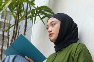 ritratto di attraente asiatico hijab donna lettura libro nel balcone. musulmano ragazza godendo gratuito volta. concetto di alfabetizzazione e conoscenza foto