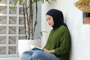 ritratto di attraente asiatico hijab donna lettura libro nel balcone. musulmano ragazza godendo gratuito volta. concetto di alfabetizzazione e conoscenza foto