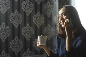 donna asiatica sorridente che beve caffè mentre usa un telefono cellulare per lavoro foto