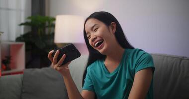 ritratto di giovane asiatico donna gode giocando in linea esport Giochi su smartphone seduta su divano nel il vivente camera a casa, giocatore stile di vita concetto. foto