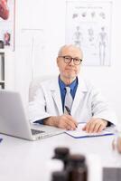 maturo medico nel ospedale ufficio indossare laboratorio cappotto mentre guardare a telecamera. medico professionista utilizzando taccuino nel clinica posto di lavoro , fiducioso, competenza, medicinale. foto