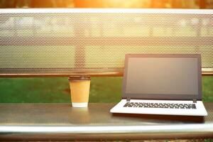 modello Immagine di il computer portatile con vuoto nero schermo e caffè tazza su metallo panchina nel natura all'aperto parco foto