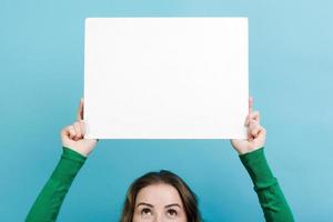 ragazza del primo piano che tiene uno spazio della copia del bordo bianco sopra la sua testa contro il fondo blu foto