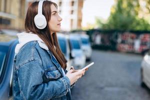 ragazza su uno sfondo urbano, in cuffie bianche e uno smartphone in mano, appoggiata all'auto foto