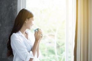 donne asiatiche che bevono caffè e si svegliano nel suo letto completamente riposate e aprono le tende al mattino per prendere aria fresca al sole foto