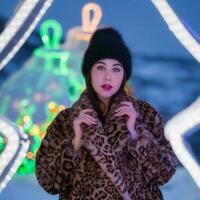 ritratto di giovane donna nel nero cappello e pelliccia cappotto con leopardo modello sta su sfondo di Natale luci decorazioni e guardare a telecamera. profondità di campo, selettivo morbido messa a fuoco su primo piano foto