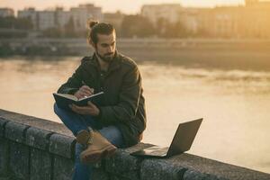 bello uomo d'affari utilizzando personale organizzatore e il computer portatile mentre seduta di il fiume.tono Immagine foto