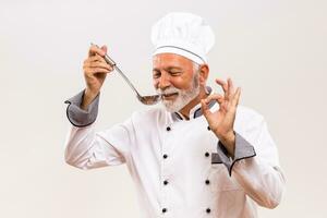 ritratto di soddisfare anziano capocuoco degustazione la minestra e mostrando ok cartello. foto