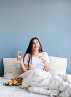 giovane donna bruna seduta sveglia nel letto con palloncini e decorazioni a forma di cuore rosso bevendo champagne mangiando croissant