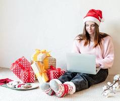 giovane donna con cappello da Babbo Natale che fa shopping online circondata da regali