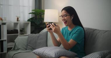 ritratto di giovane asiatico donna indossare bicchieri e cuffie gode giocando in linea esport Giochi su smartphone seduta su divano nel il vivente camera a casa, giocatore stile di vita concetto. foto
