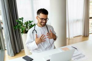 medico con il computer portatile conduce un in linea video chiamata e parla per paziente. maschio medico Lavorando a ufficio scrivania, ufficio interno su sfondo foto