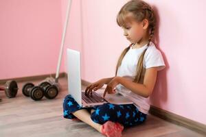 carino adorabile caucasico poco biondo ragazza seduta durante in linea video Chiacchierare scuola lezione sessione con insegnante e classe. a distanza formazione scolastica concetto. auto-isolamento a quarantena foto