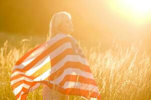 bellissimo giovane ragazza Tenere un americano bandiera nel il vento nel un' campo di segale. estate paesaggio contro il blu cielo. orizzontale orientamento. foto