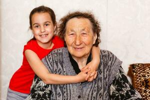 famiglia bonding concetto. bello poco ragazza felicemente abbracciare sua nonna nel leggero vivente camera foto