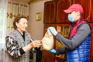un anziano donna soggiorni a casa. cibo consegna nel un' medico maschera per il anziano. foto