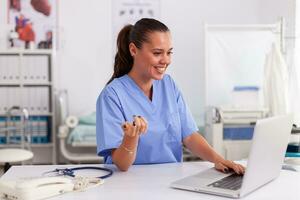 medico infermiera sorridente mentre utilizzando il computer portatile nel ospedale ufficio indossare blu uniforme. Salute cura professionista seduta a scrivania utilizzando computer nel moderno clinica guardare a tenere sotto controllo, medicinale. foto