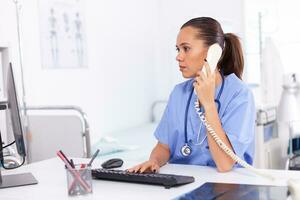 medico infermiera parlando con paziente su il Telefono di diagnosi. Salute cura medico seduta a scrivania utilizzando computer nel moderno clinica guardare a tenere sotto controllo. foto