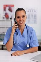 medico personale parlando con paziente su Telefono a partire dal ospedale ufficio di diagnosi sorridente a telecamera. femmina infermiera, medico avendo un' Telefono conversazione con malato persona durante consultazione, medicinale. foto