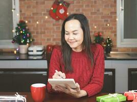 asiatico donna indossare rosso a maglia maglione seduta a tavolo con rosso tazza di caffè e regalo scatole nel il cucina con Natale decorazione, utilizzando penna scrittura nome elenco su taccuino. foto