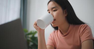 ritratto giovane asiatico donna digitando al di sopra di il tastiera di sua il computer portatile mentre seduta bevanda caffè nel vivente camera a domestico, sociale networking foto