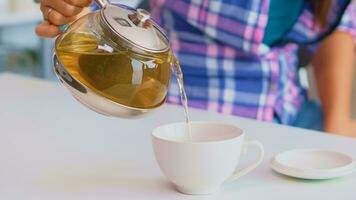 vicino su di tè a partire dal il bollitore lentamente versare in porcellana tazza. giovane signora preparazione verde tè nel il cucina nel il mattina a colazione, utilizzando teiera, tazza di tè e sano erbaceo le foglie. foto