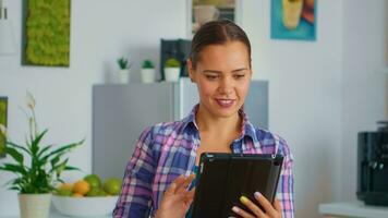 donna utilizzando tavoletta seduta a il tavolo nel il cucina. signora sorridente lettura notizia su tavoletta mentre potabile verde tè Lavorando a partire dal casa utilizzando dispositivo con Internet tecnologia digitando, navigazione su gadget. foto