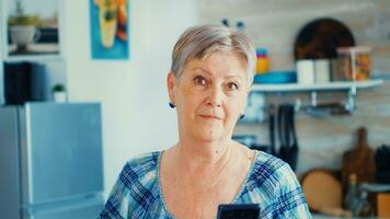 nonna guardare a telecamera mentre utilizzando smartphone nel cucina durante prima colazione. autentico anziano persona utilizzando moderno smartphone Internet tecnologia. in linea comunicazione collegato per il mondo, anziano tempo libero tempo con aggeggio a la pensione età foto