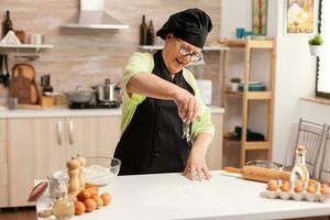 allegro anziano signora fabbricazione Pizza nel casa cucina utilizzando superiore Farina per cottura al forno. contento anziano capocuoco con uniforme aspersione, setacciare vagliatura crudo ingredienti di mano. foto