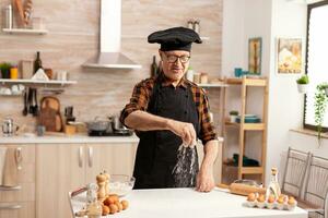 anziano panettiere preparazione pane utilizzando bio Grano Farina nel casa cucina indossare grembiule e bonete. cucinare nel cucina uniforme aspersione setacciare vagliatura ingredienti di mano. foto